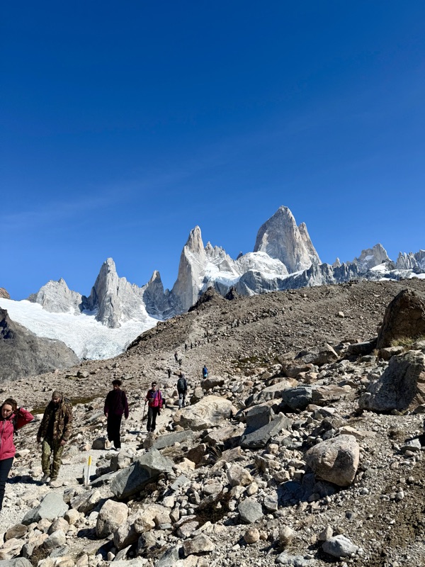 Hikers on a rocky trail climbing towards the jagged, snow-flecked peaks of Fitz Roy under a deep blue sky