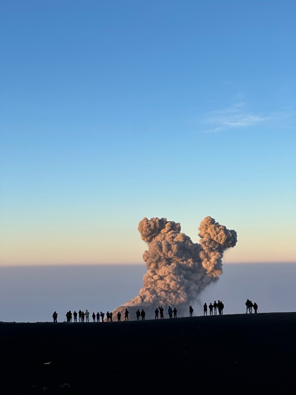 A tall ash cloud rising from Volcán de Fuego above a sea of clouds, a line of people silhouetted on the ridge in front