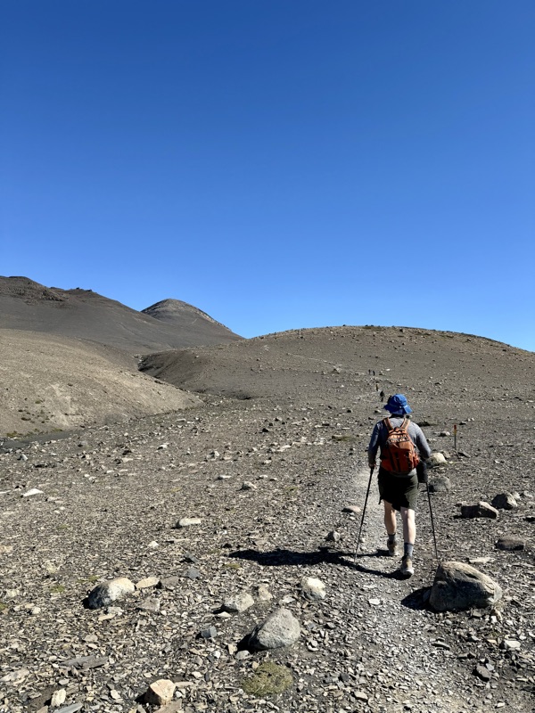 A hiker with trekking poles and an orange backpack walking a stony path across a bare ridge under a cloudless sky