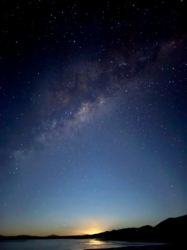 The Milky Way arching over a still lagoon, the moon rising in an orange glow behind dark hills