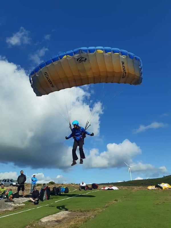 Lifting off under a blue and yellow paraglider from a grassy launch, wind turbines on the ridge behind