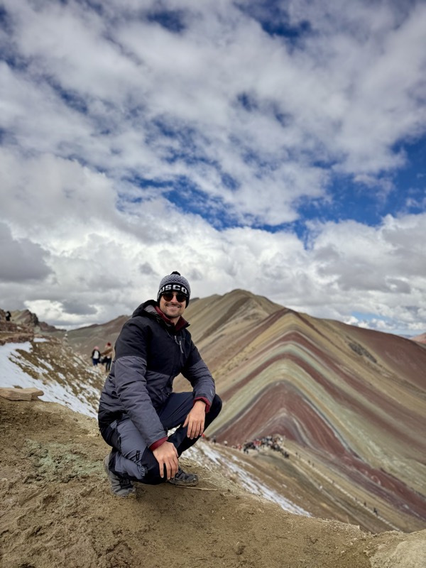 Crouching in a beanie and down jacket on a dusty ridge, the red, ochre and green stripes of Rainbow Mountain behind under a cloudy sky