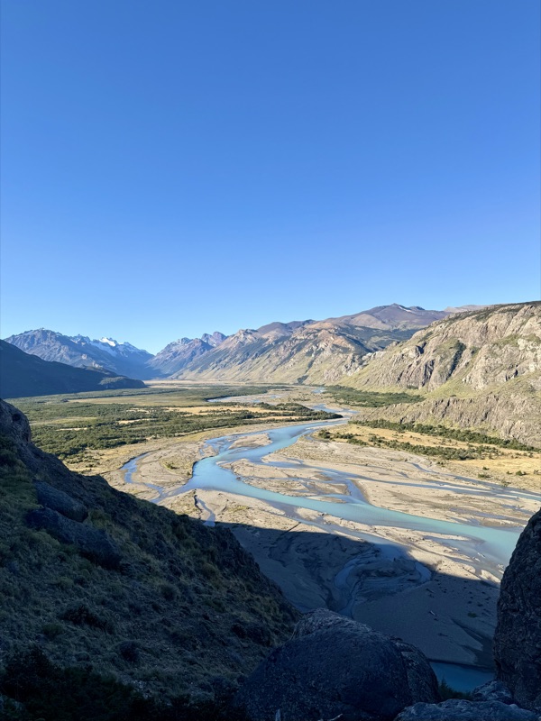 A braided turquoise river winding along a wide valley floor between dry mountains, seen from a rocky slope above