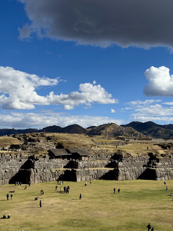 The zigzag stone walls of Sacsayhuamán along a green esplanade above Cusco, under big clouds