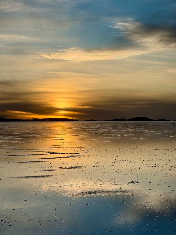 Golden sunset mirrored in a thin layer of water on the Salar de Uyuni, low hills on the horizon