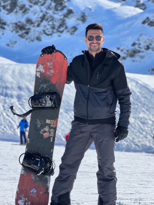 Smiling in sunglasses, holding a red snowboard upright on a sunny snow slope